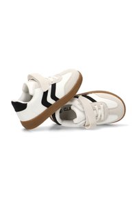 Zapatillas blancas deportivas con acentos negros, que cuentan con una correa de velcro, una suela acolchada y un upper liso similar al cuero, junto con una suela exterior texturizada de color marrón.