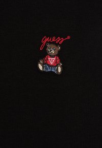 Melnā auduma, uz kura ir izšūts lācis sarkanā krekliņā ar baltu logotipu, kopā ar sarkano paliekamo tekstu "guess."