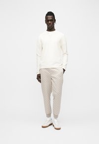 Mees seisab, kandes elevandiluurõngast pikkade varrukatega henley-t, musteriga beeže pükse ja valgeid tossusid kummist tallaga.