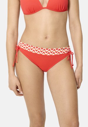 Mujer con bikini rojo con cinturilla estampada y lazos ajustables en los lados de la parte inferior del bikini contra un fondo liso.