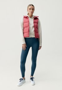 Röd puffig väst över en beige hoodie, kombinerad med mörkblå leggings och vita sneakers. Vesten har dragkedja och huva.