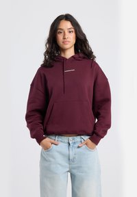 Young woman with long dark hair wearing a maroon hoodie with "The Botanical Garden" text and light blue jeans, hands in pockets.