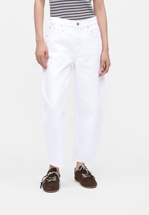 Person iført hvide jeans med lige ben, stribet ærmeløs top og brune snøresko mod en ensfarvet hvid baggrund.