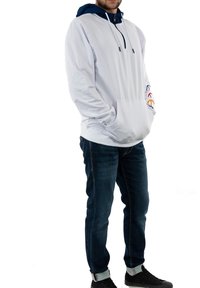 Hombre vistiendo una sudadera con capucha blanca con logotipos multicolores en las mangas, jeans oscuros y zapatillas negras, de pie con las manos en los bolsillos.