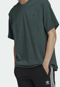 Eine Person trägt ein dunkelgrünes, kurzärmliges T-Shirt und schwarze Adidas-Jogginghosen mit weißen Streifen, die Hände in den Taschen, in einer lässigen Pose.