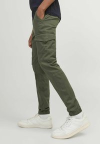 Pantalons cargo en vert olive, fabriqués en tissu coton, avec des poignets élastiques, plusieurs poches latérales et une coupe décontractée. Baskets blanches visibles.