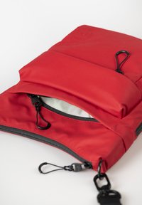 Borsa in tessuto morbido rosso con patta superiore, cerniera nera e tracolla regolabile. L'interno presenta una fodera chiara e una tasca di stoccaggio.