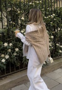 Per didelis baltas dryžuotas marškinis, šviesiai rudas megztinis, užmestas ant pečių, baltos plačios kelnės, laikantis ledinį gėrimą, žali lapai fone.