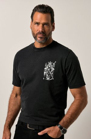 Camiseta de algodón negra con un diseño gráfico blanco en el lado izquierdo del pecho, mangas cortas y un cuello acanalado. Llevada con pantalones negros y un reloj.