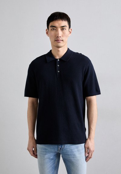 Marineblå polo-shirt med et struktureret strikdesign, tre knaplukninger, korte ærmer og en lige kant, parret med lyseblå jeans.