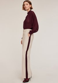 Bordeaux doorschijnende blouse met een hoge hals en lange mouwen, gecombineerd met crèmekleurige broeken met een donker bordeaux zijstreep.