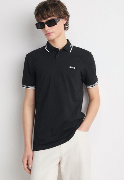Polo noir avec col et ourlets de manches à rayures blanches ; présente un petit logo blanc sur la poitrine ; porté avec un pantalon de couleur claire.