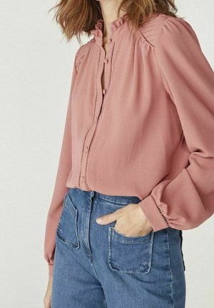 Kvinde iført en pink langærmet bluse med knapper og flæset krave samt blå højtaljede jeans, hånd i lommen.