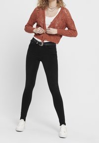 Mujer abrochándose un cárdigan de punto color óxido sobre una camiseta blanca, vistiendo jeans skinny negros, zapatillas blancas y collares en capas.
