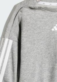 Grå sweatshirt med en mjuk textur, med vita tre-randsdetaljer på ärmarna och en liten logotyp på framsidan.