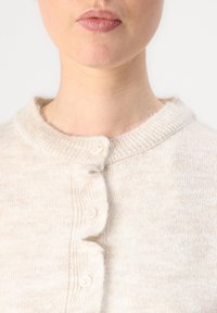 Cárdigan abotonado en color beige claro, confeccionado en una suave tela texturizada. Presenta un cuello de canalé y cuatro botones a lo largo de la parte delantera.