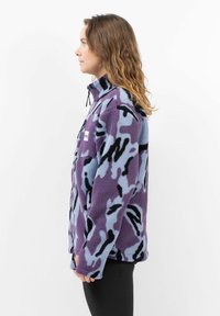 Fleecejack in paars en lichtblauw met een zwart abstract patroon. Bevat een ritskraag, lange mouwen en een zak op de borst.