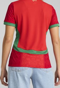 Widok z tyłu osoby w czerwono-zielonym sportowym podkoszulku w wzory oraz jasnoniebieskich dżinsach, stojącej z rękami swobodnie wzdłuż ciała.