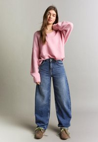 Tânără cu păr lung, purtând un pulover roz supradimensionat, blugi largi albaștri și adidași maro cu șireturi verzi, stând în fața unui fundal simplu.