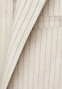 Tissu de veste de costume beige à fines rayures verticales noires étroites avec une petite poche poitrine visible.