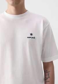 T-shirt en coton blanc avec un col rond et des manches courtes, ornée d'un petit logo brodé noir indiquant "snow peak" sur la poitrine.