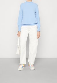 Top azzurro chiaro a maniche lunghe con scollatura arricciata, abbinato a pantaloni bianchi larghi e sneakers multicolori, mentre tiene una piccola borsa bianca.