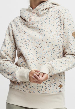 Kobieta w kremowej bluzie z kapturem z niebieskim i brązowym wzorem kropek, trzema guzikami przy szyi oraz ściągaczami przy mankietach i dolnej krawędzi.