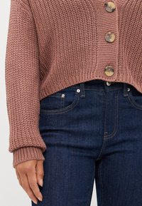 Suéter marrón de punto con un diseño corto, botones grandes y puños acanalados, combinado con jeans de mezclilla oscuros que tienen bolsillos laterales.