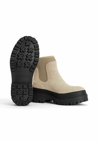 Beige ankelstøvler med nubuck-tekstur, elastiske sidelapper og en tyk sort gummisål med et mønster for bedre greb.
