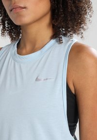 Top senza maniche Nike azzurro chiaro con collo tondo, caratterizzato da un logo argento. Tessuto morbido e vestibilità rilassata, indossato sopra un top nero.