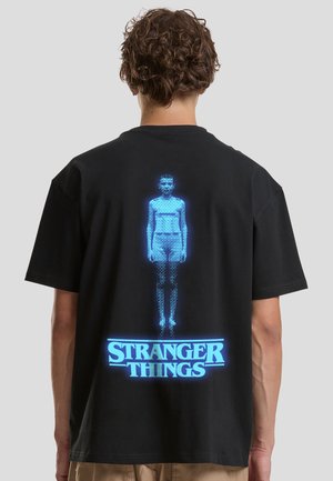 Tricou negru din bumbac cu un grafic albastru reprezentând o figură și textul „STRANGER THINGS” dedesubt. Mâneci scurte și croială lejeră.