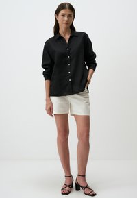 Camicia button-up nera in lino a maniche lunghe, abbinata a pantaloni corti di colore chiaro e sandali con tacco alto neri e cinturini.