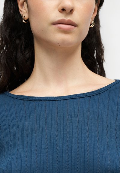 Gros plan sur le bas du visage et le cou d’une personne portant des boucles d’oreilles anneaux dorés et un haut bleu côtelé à encolure bateau.