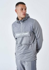 Jacket grigia leggera con cappuccio e tasca frontale con logo. Presenta dettagli con zip e cordini regolabili attorno al cappuccio.