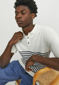 Hvid polo shirt med navy horisontale striber, korte ærmer og tre-knaps knaplukning. Model bærer blå denimbukser og sidder på en træstol.
