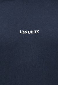 Ύφασμα ναυτικού μπλε με λευκό κεντητό κείμενο "LES DEUX" στο κέντρο της επιφάνειας.