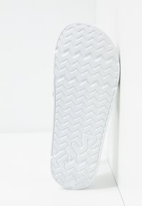 Semelle d'une chaussure blanche sans lacets avec un motif en zigzag et le nom de la marque embossé, positionnée à la verticale contre un mur d'angle blanc.