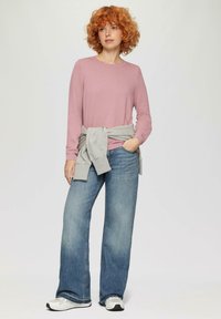 Roze textuur lange mouwen top, grijze sweatshirt om de taille geknoopt, blauwe wijde jeans en witte sneakers.