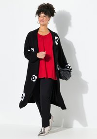 Langer schwarzer Cardigan mit weißen Blumenakzenten, getragen über einer roten Bluse und schwarzen Hosen. Verfügt über eine weiche Textur und eine lockere Passform. Silbergraue Knöchelstiefel.