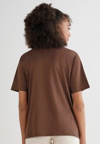 T-shirt in cotone marrone con una vestibilità rilassata, maniche corte e una trama liscia. La vista posteriore mostra un collo pulito e cuciture minime.