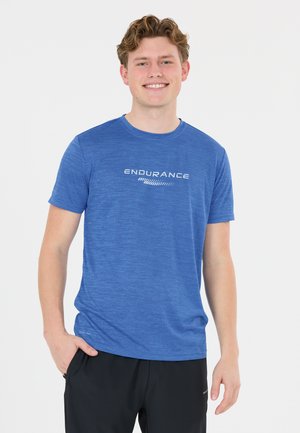 Lächelnder junger Mann mit lockigem Haar, der ein blaues "Endurance"-T-Shirt und schwarze Hose trägt und mit einer Hand in der Tasche steht.