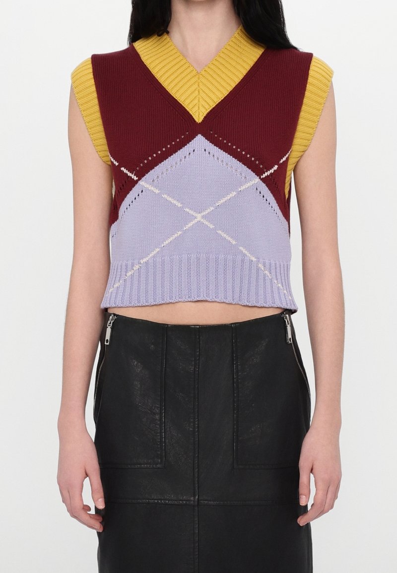 Gilet en tricot sans manches avec motifs géométriques bordeaux, lavande et moutarde, porté avec une jupe en cuir noire taille haute avec fermetures éclair latérales.