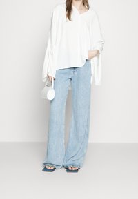 Top bianco largo con maniche lunghe e scollatura a V, abbinato a jeans azzurri a gamba larga e sandali blu scuro con apertura. Piccola borsa bianca.
