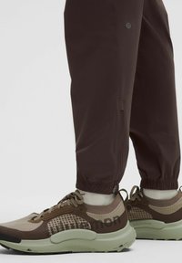 Braune Sporthosen verfügen über einen elastischen Bund und einen Reißverschluss. Die Sneaker sind beige mit braunen Akzenten und haben ein strukturiertes Muster auf der Oberseite.