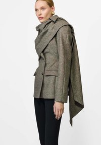 Podwójnie zapinana marynarka z teksturowanej tkaniny w odcieniach szaro-brązowych, z dużym detalem szala zarzuconym na jedno ramię oraz dopasowaną sylwetką.