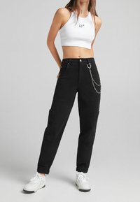 Svarta cargo-byxor med ribbade mudd, flera fickor och en metallkedjeaccent fäst vid en bälteloop. Bärs tillsammans med en vit crop top.