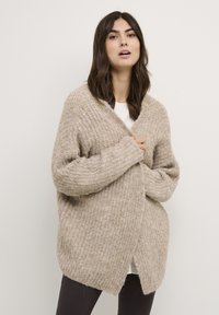 Gilet en tricot beige à texture côtelée, à l'avant ouvert et à manches longues. Présente une coupe décontractée et des fentes sur les côtés. Porté par-dessus un haut blanc.