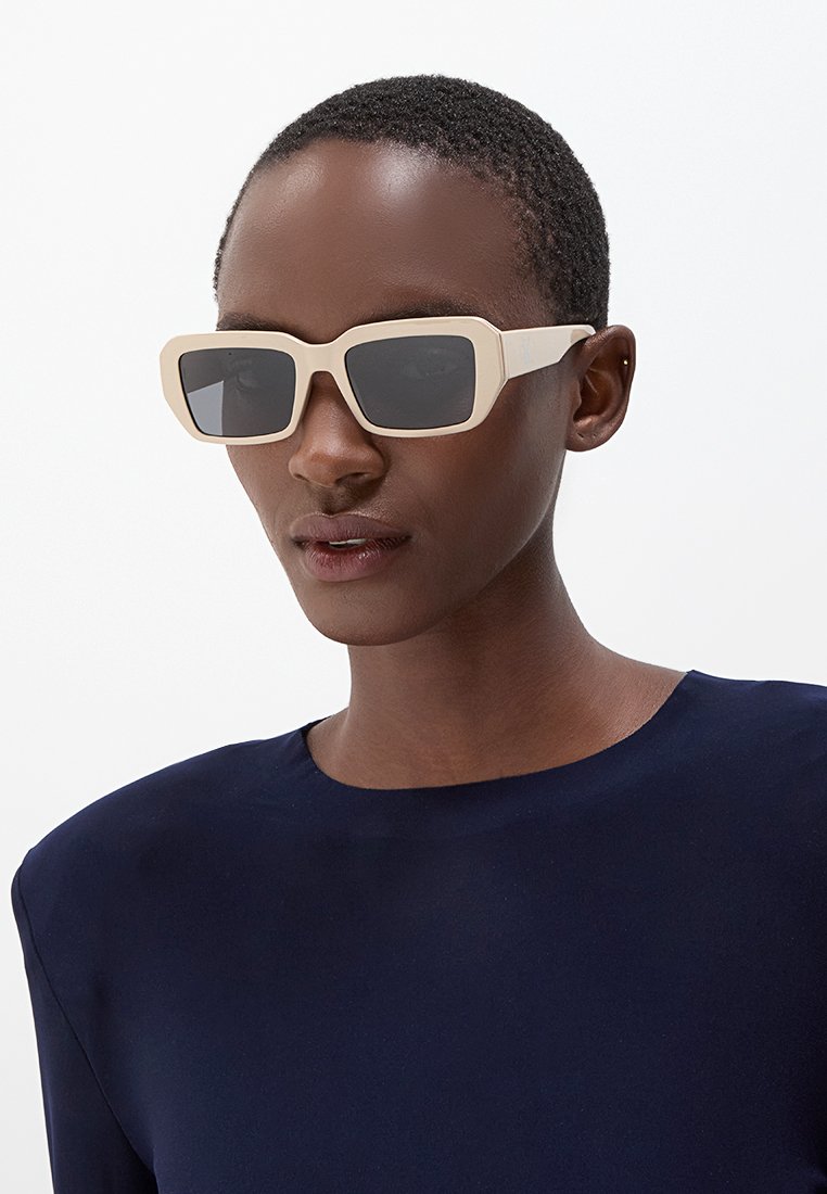 Kwadratowe okulary przeciwsłoneczne z grubą, beżową plastikową oprawą i ciemnoszarymi soczewkami. Nosi je osoba w granatowej bluzce, z krótkimi, bardzo przystrzyżonymi włosami.