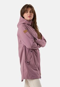 Žena v hlohovo fialovom pršiplášti s kapucňou a prednými vreckami, stojaca so založenými rukami a pozorujúca mierne do bokua na jednotnom pozadí.