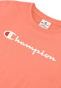 Camiseta color coral con cuello redondo. Presenta un logo blanco de "Champion" en texto cursivo en la parte delantera. Fabricada en suave material de algodón.